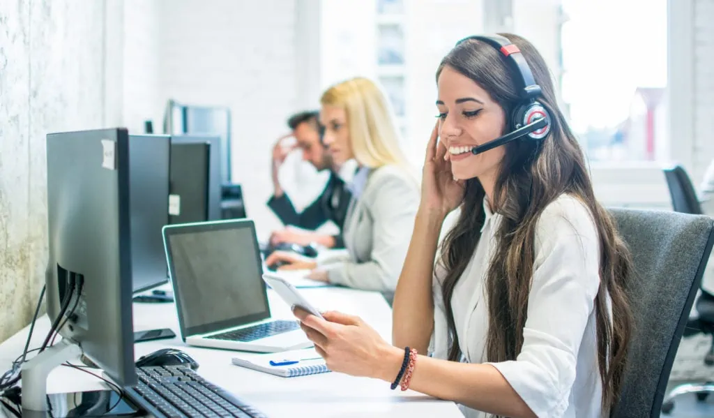 Friendly female customer support operator with headset using phone in office