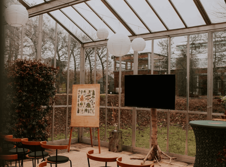 Binnen in Het Groene Huis, een lichte kas met hoge tafels, krukken, een beeldscherm en lampionnen